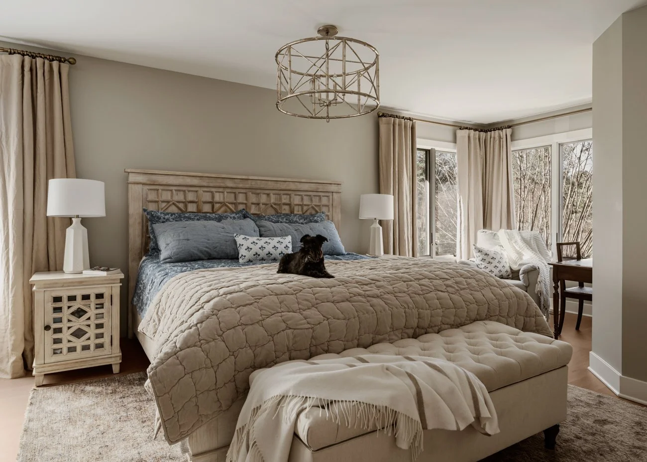 Bedroom with a large bed, nightstands with lamps, a bench at the foot of the bed, and windows with curtains revealing a view of trees outside. A small dog sits on the bed.