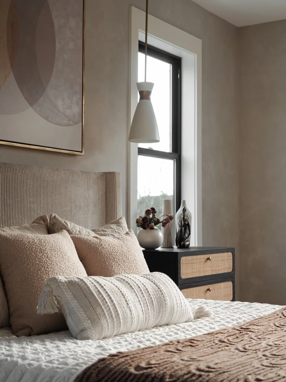 A cozy bedroom with a bed, beige pillows, and a textured cream pillow in front. There is a black nightstand with a woven drawer front, holding a vase of flowers and glass decorative objects. A window allows natural light, and a modern hanging lamp is