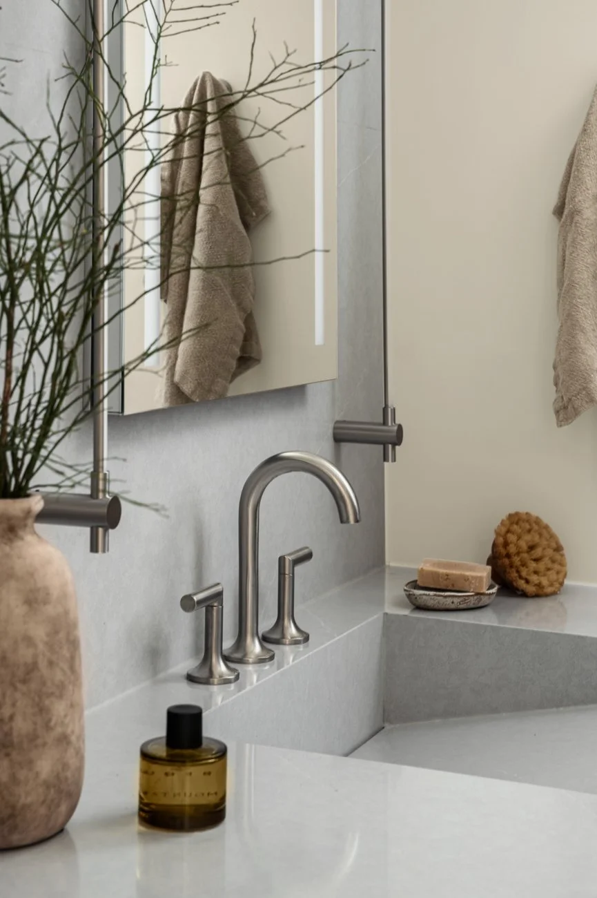 A bathroom vanity with a beige towel hanging on a wall, a mirror, a soap dish with soap, a decorative sponge, a bar of soap, a bottle of facial serum, and a vase with branches.