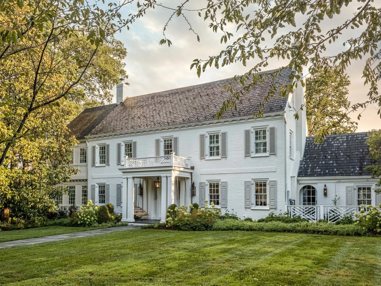 A large white house with gray shutters and a gray shingle roof, surrounded by a manicured lawn and trees with autumn leaves.