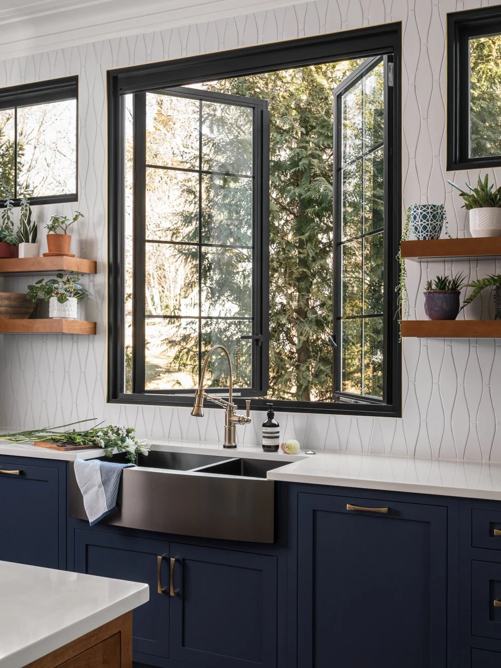 Kitchen with open window, white patterned backsplash, blue cabinets, and open wooden shelves with potted plants and decorative items.