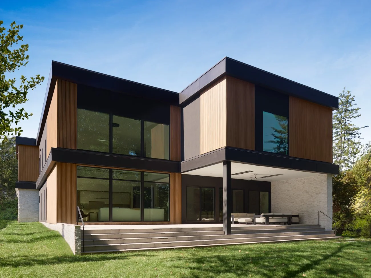 Modern two-story house with large windows, wooden and white exterior, set in a green grassy yard, under a clear blue sky.