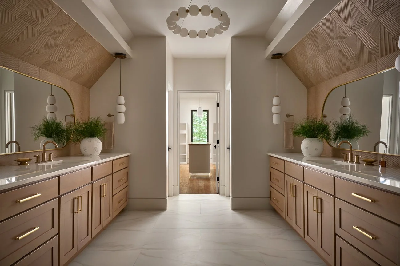 A modern bathroom with two beige vanities with gold handles, large mirrors, pendant lights, and potted plants, leading to a room with a window in the background.