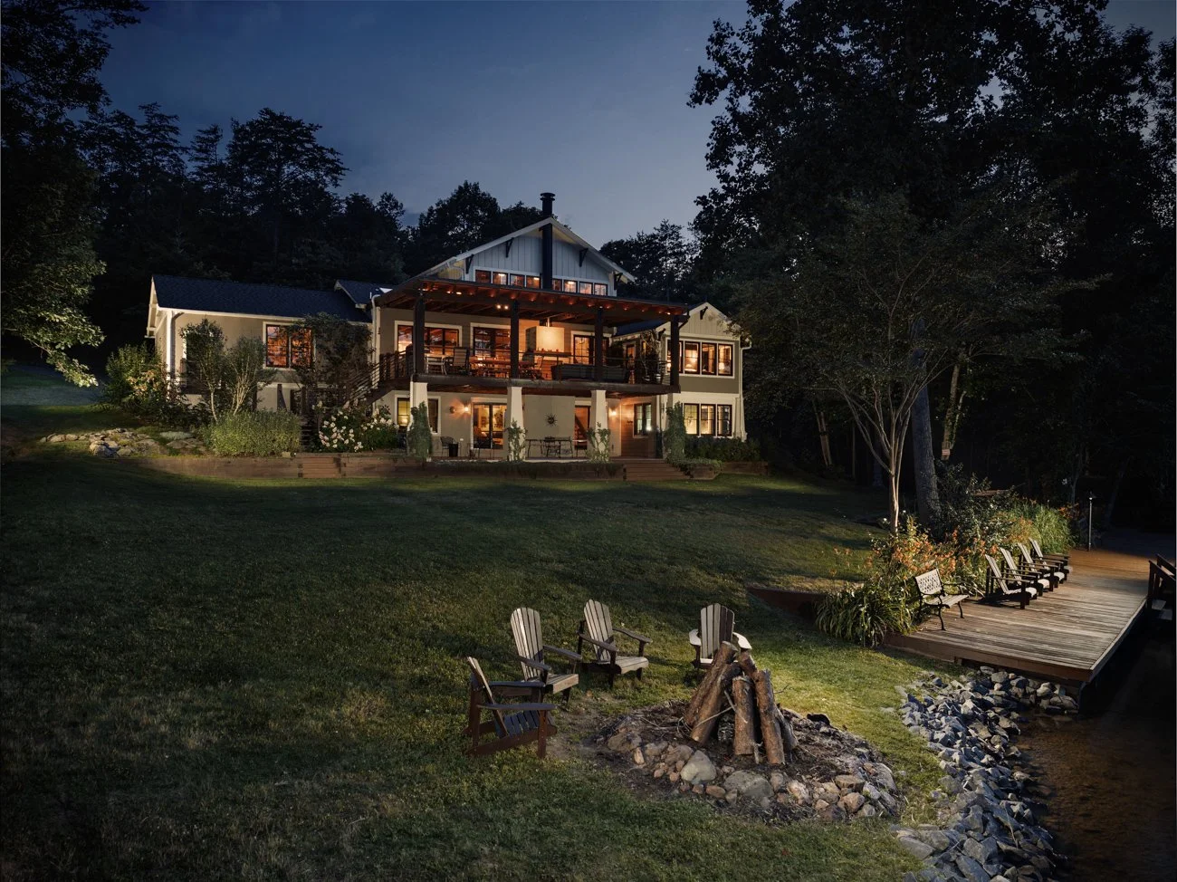 A large two-story house illuminated at night, with multiple windows and a spacious balcony, surrounded by trees and greenery, and a waterfront deck with chairs and a firepit in the foreground.