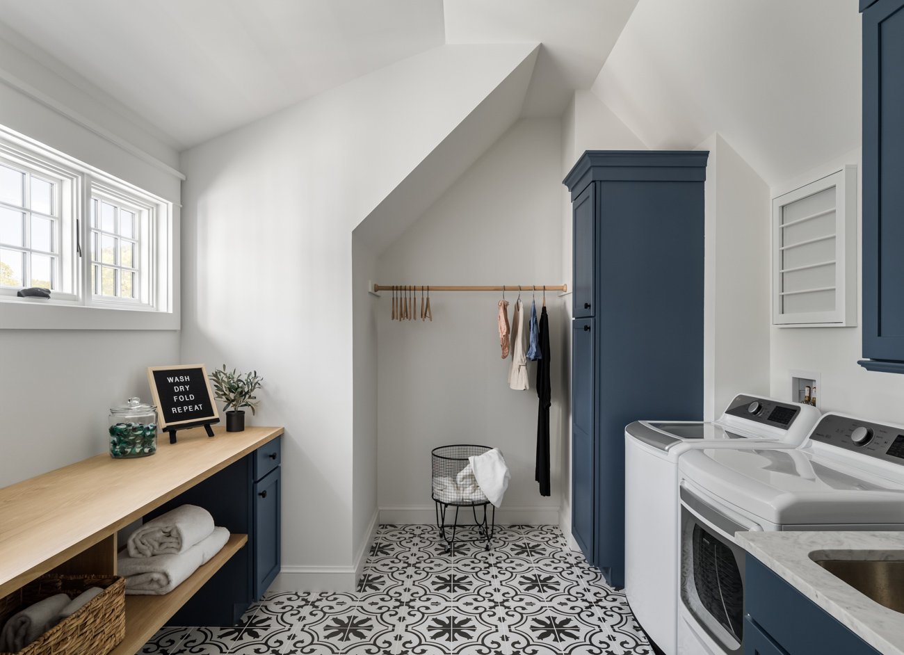 A laundry room with white walls, blue cabinets, a wooden countertop, black and white patterned floor, and a window. It features a laundry basket, a small black plant, a sign that says "Wash Dry Fold Repeat," and hanging clothes.