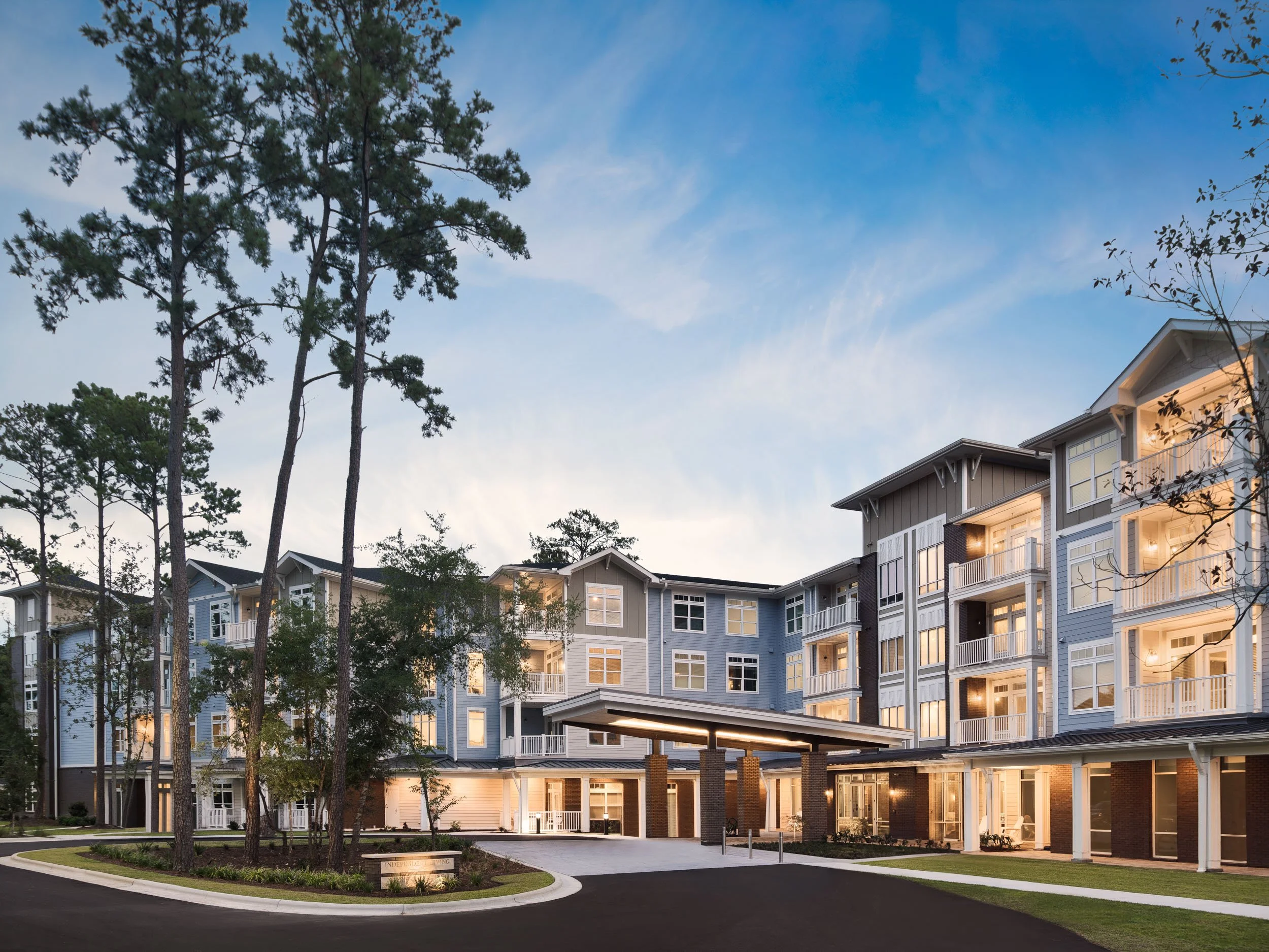 A multi-story apartment complex illuminated at dusk with trees and a curved driveway in front.
