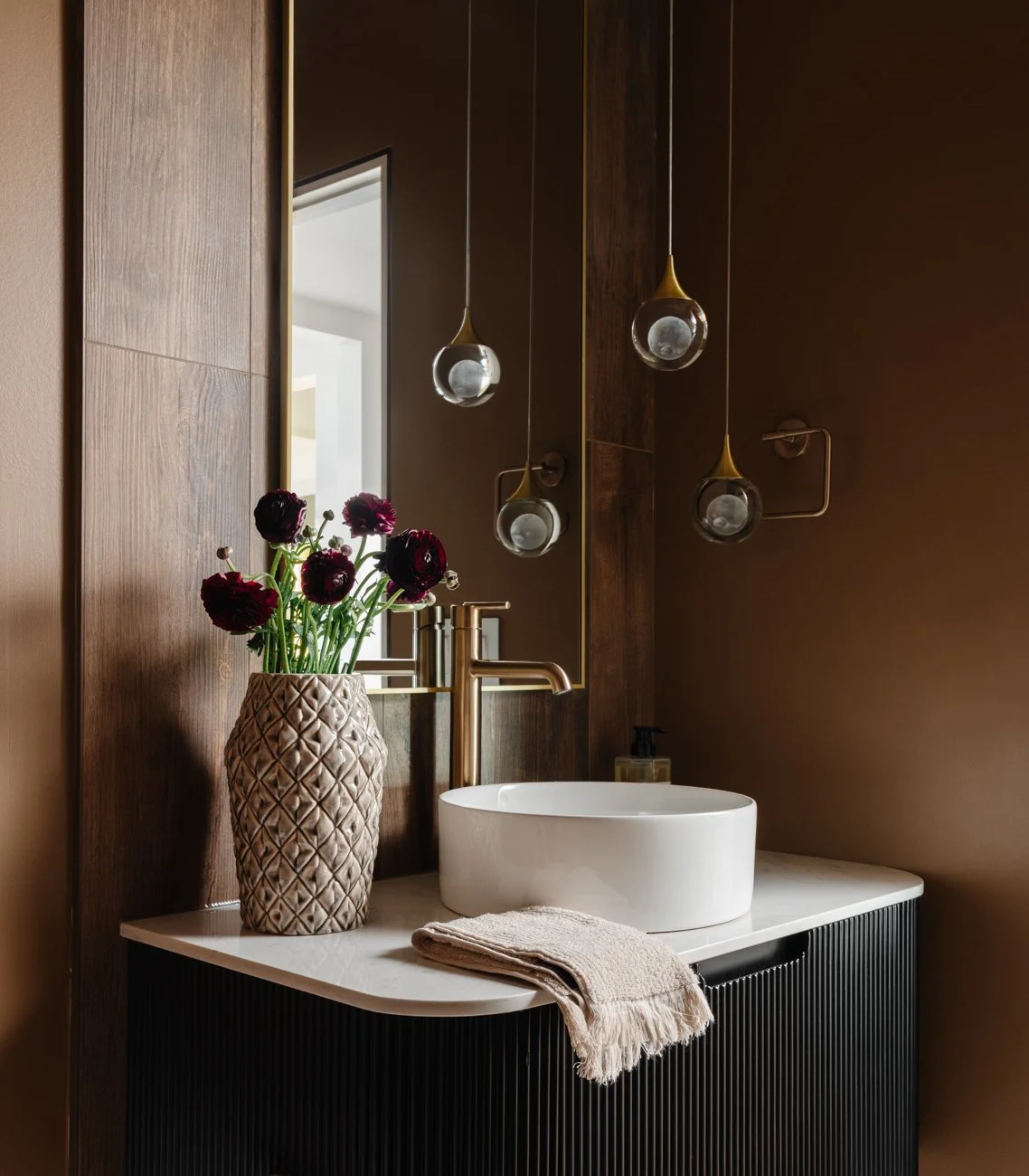 A modern bathroom vanity with a white vessel sink, gold faucet, a beige towel, and a textured beige vase with dark purple flowers, illuminated by hanging glass pendant lights.