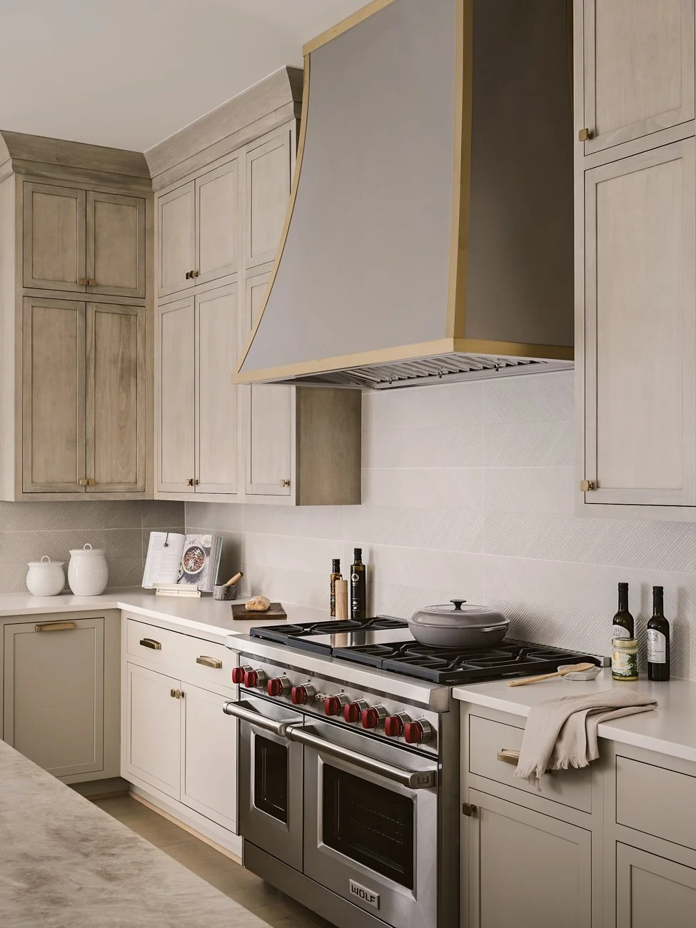 Kitchen with beige cabinets, a stove with red knobs, and a range hood. Countertop with jars, books, and bottles, and a gray cloth. Light-colored tiled backsplash.