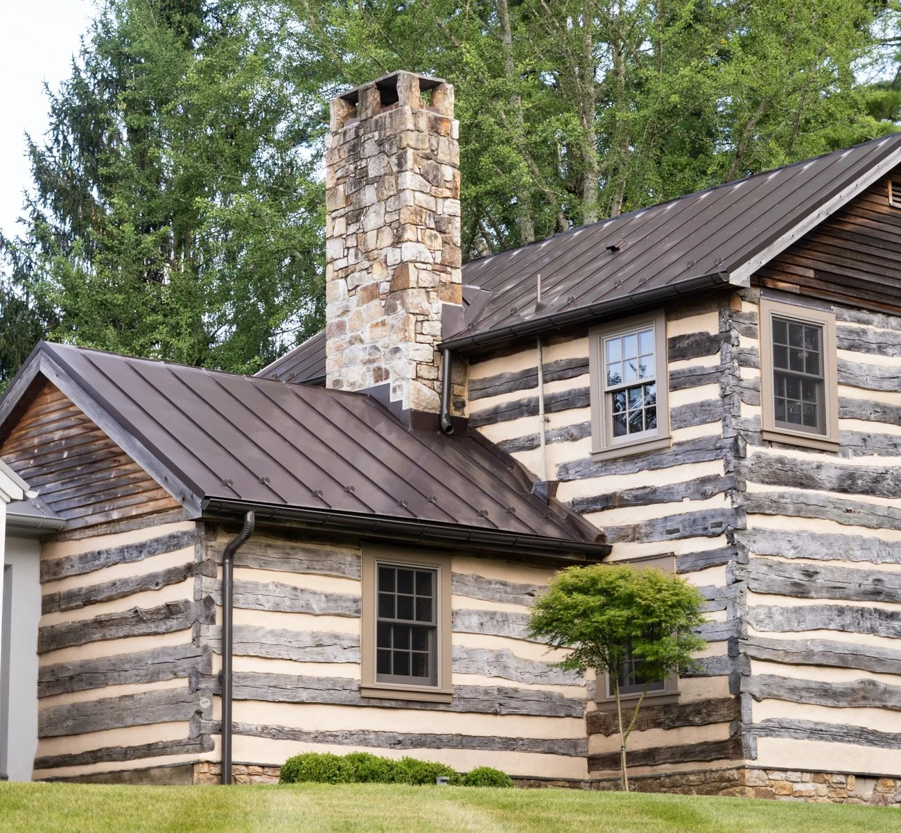 A rustic log house with a stone chimney, metal roof, and small trees in front