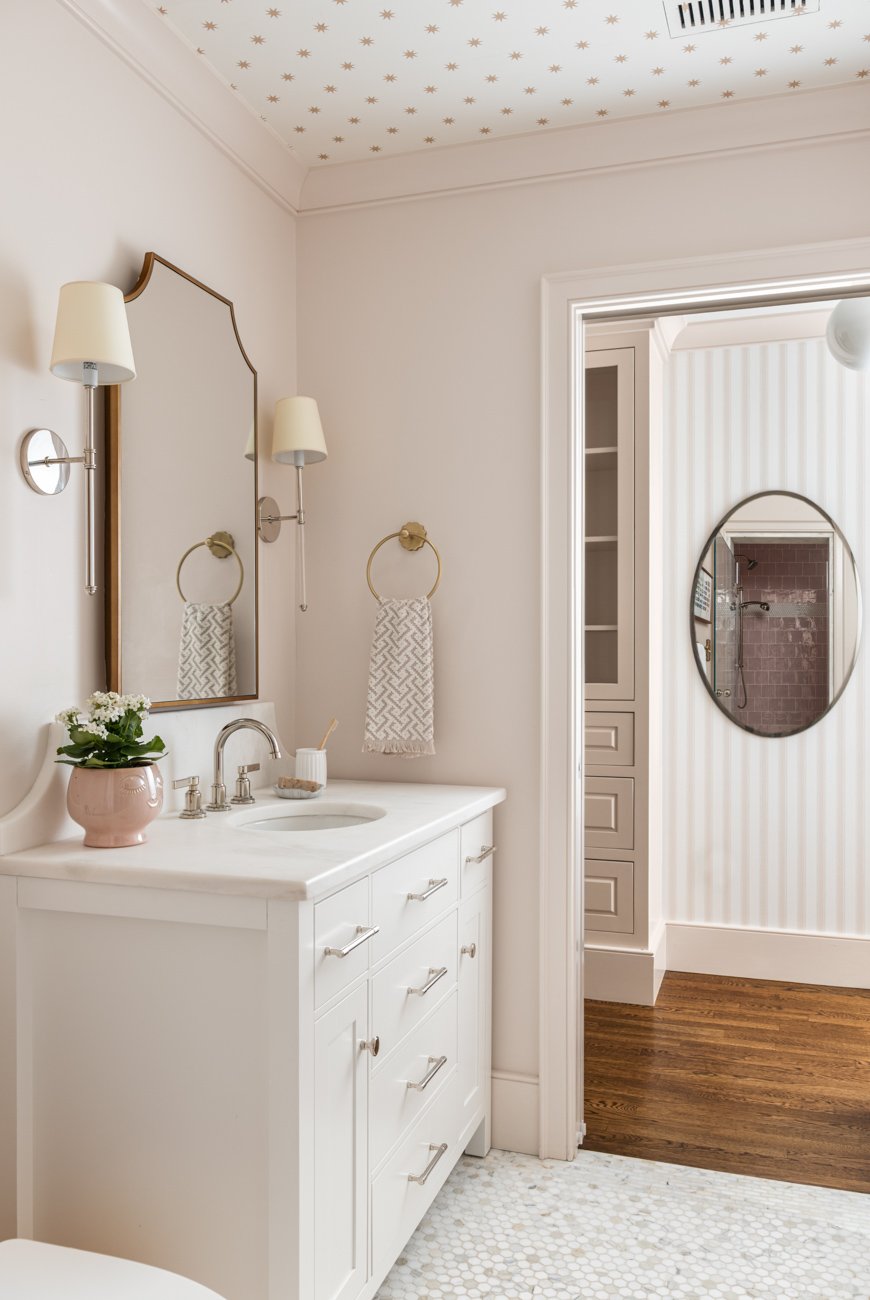 A bathroom with white walls, a white vanity with silver handles, a round mirror, wall-mounted lamps, a pink vase with white flowers, and a towel ring with a patterned towel. The ceiling has a star pattern, and there's a doorway leading to another roo