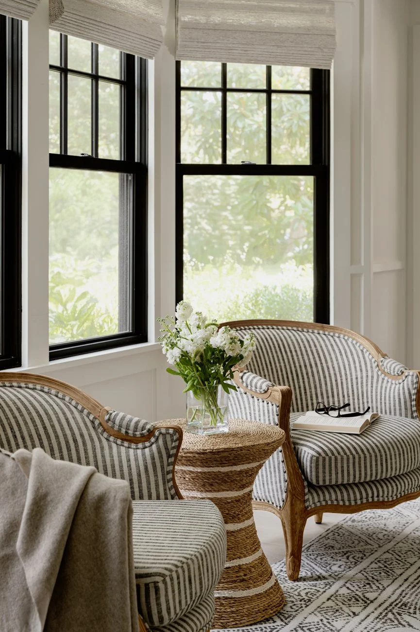 Interior of a sunlit room with three black-framed windows, striped upholstered chairs with wooden frames, a small table with a vase of white flowers, a book, and glasses on a patterned rug.