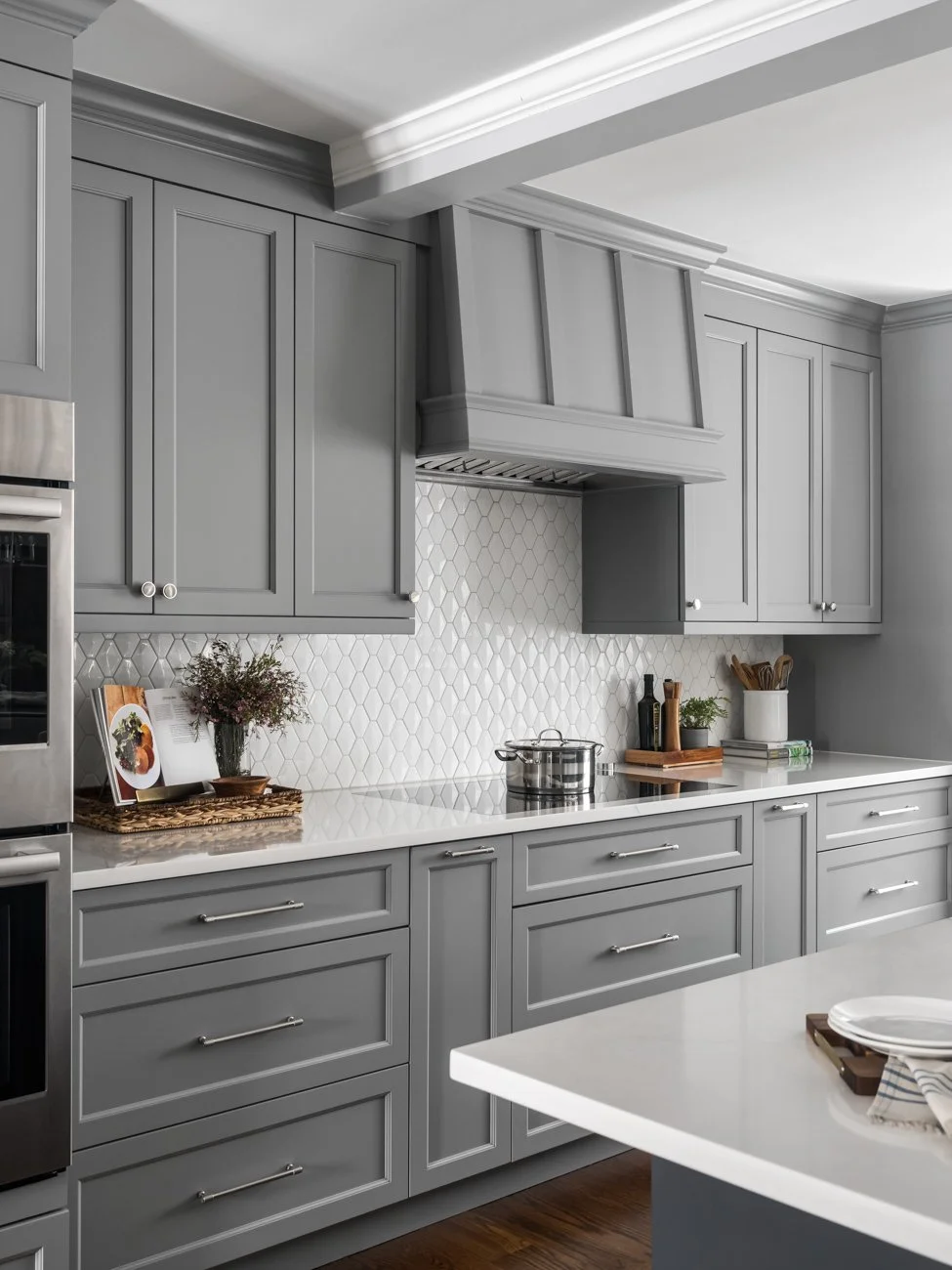 Modern kitchen with gray cabinets, white countertops, stainless steel oven, and decorative items like a plant, cookbook, and cooking utensils.