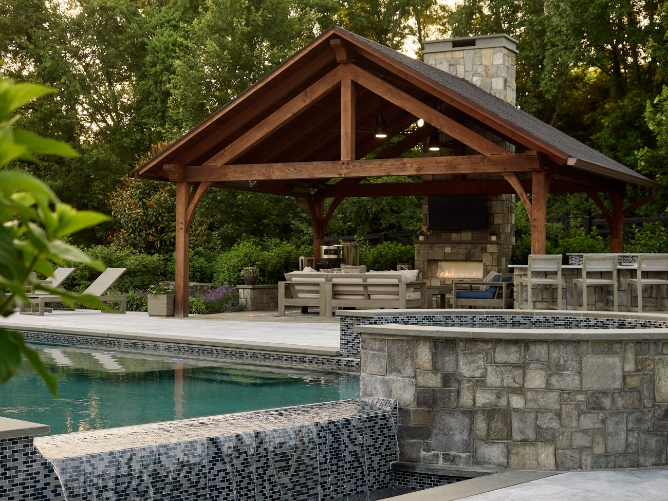Outdoor pool area with a stone fountain, wooden pavilion with a fireplace, lounge chairs, and lush greenery.