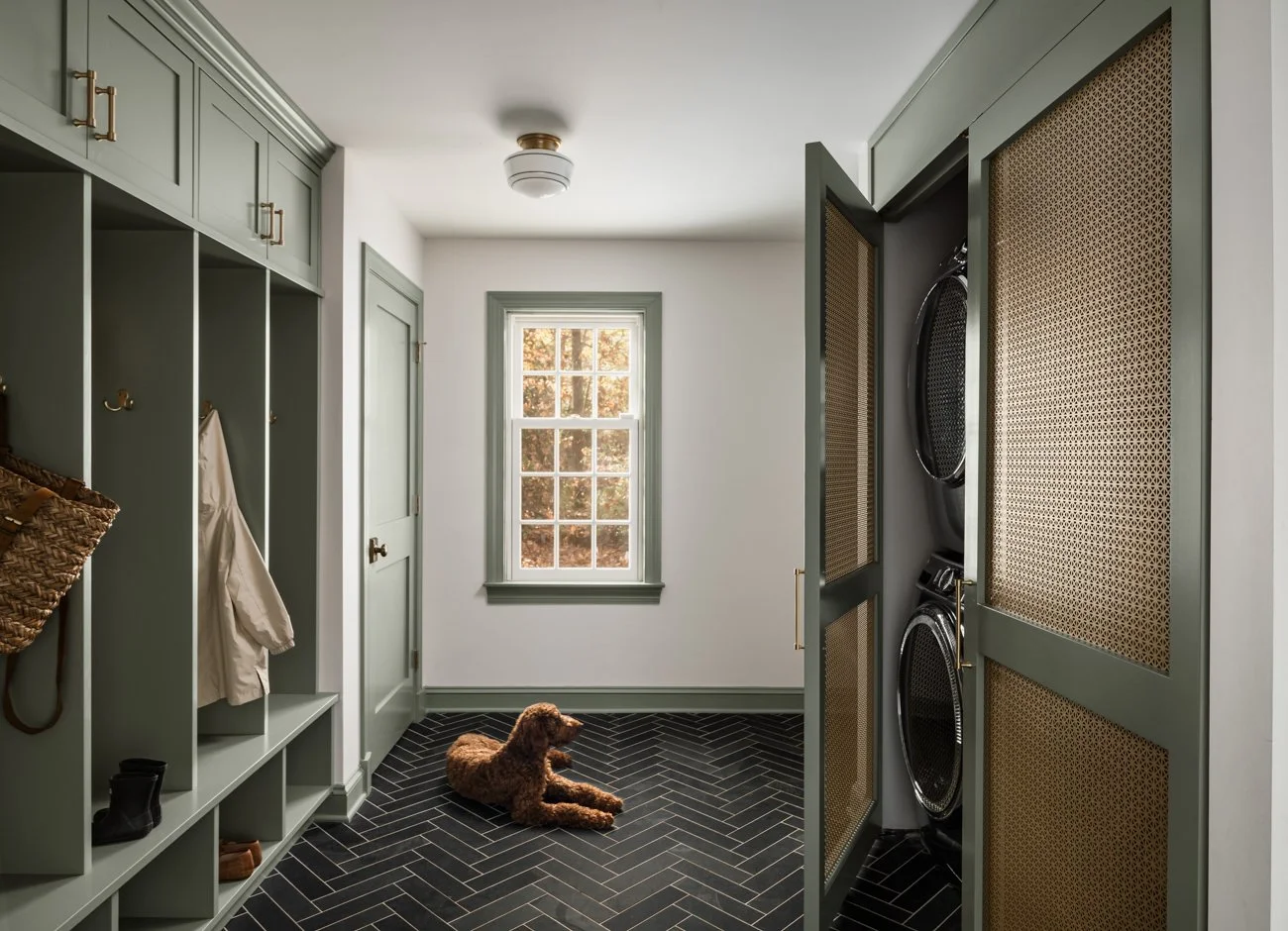Mudroom with green built-in lockers, a window showing fall foliage outside, and a washer and dryer behind a green screen door. A brown dog is lying on the black herringbone-patterned floor.