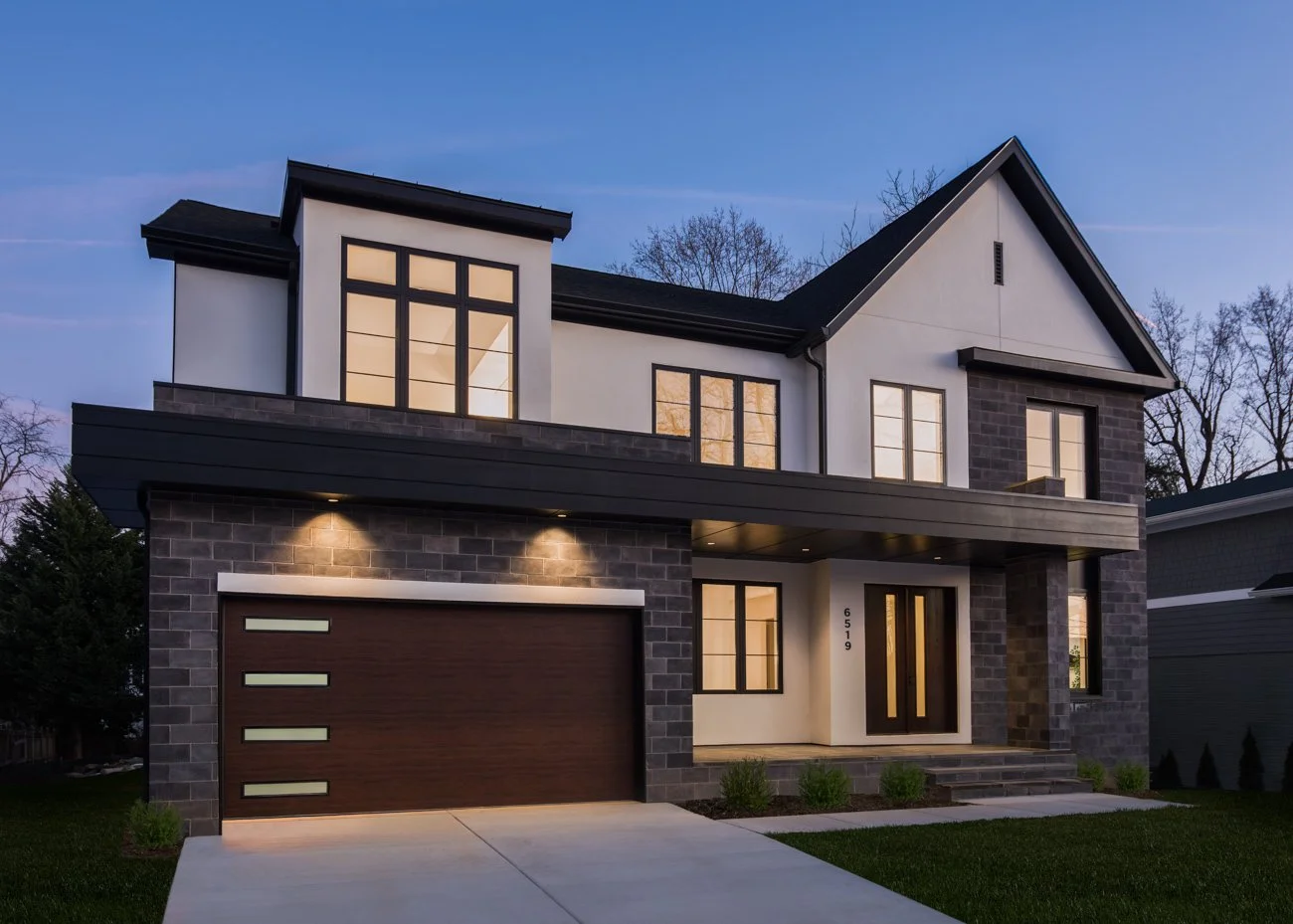 Modern two-story house with white and dark gray exterior, large windows, front steps, and a wooden garage door, illuminated at dusk.
