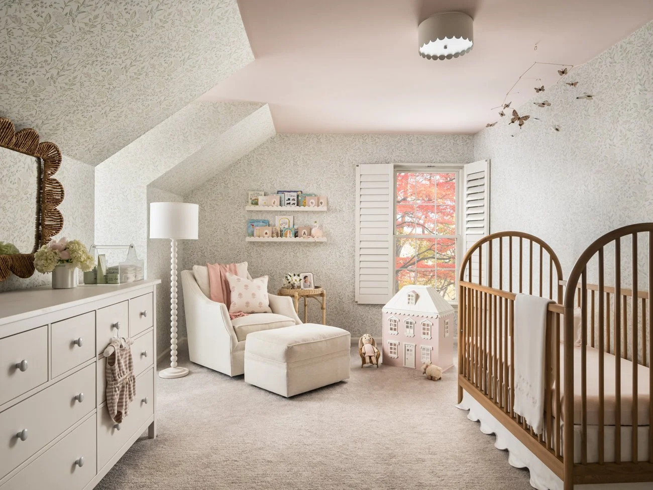 Nursery room with a crib, armchair, lamp, shelves with toys, window with autumn leaves, and decorative butterflies hanging from the ceiling.