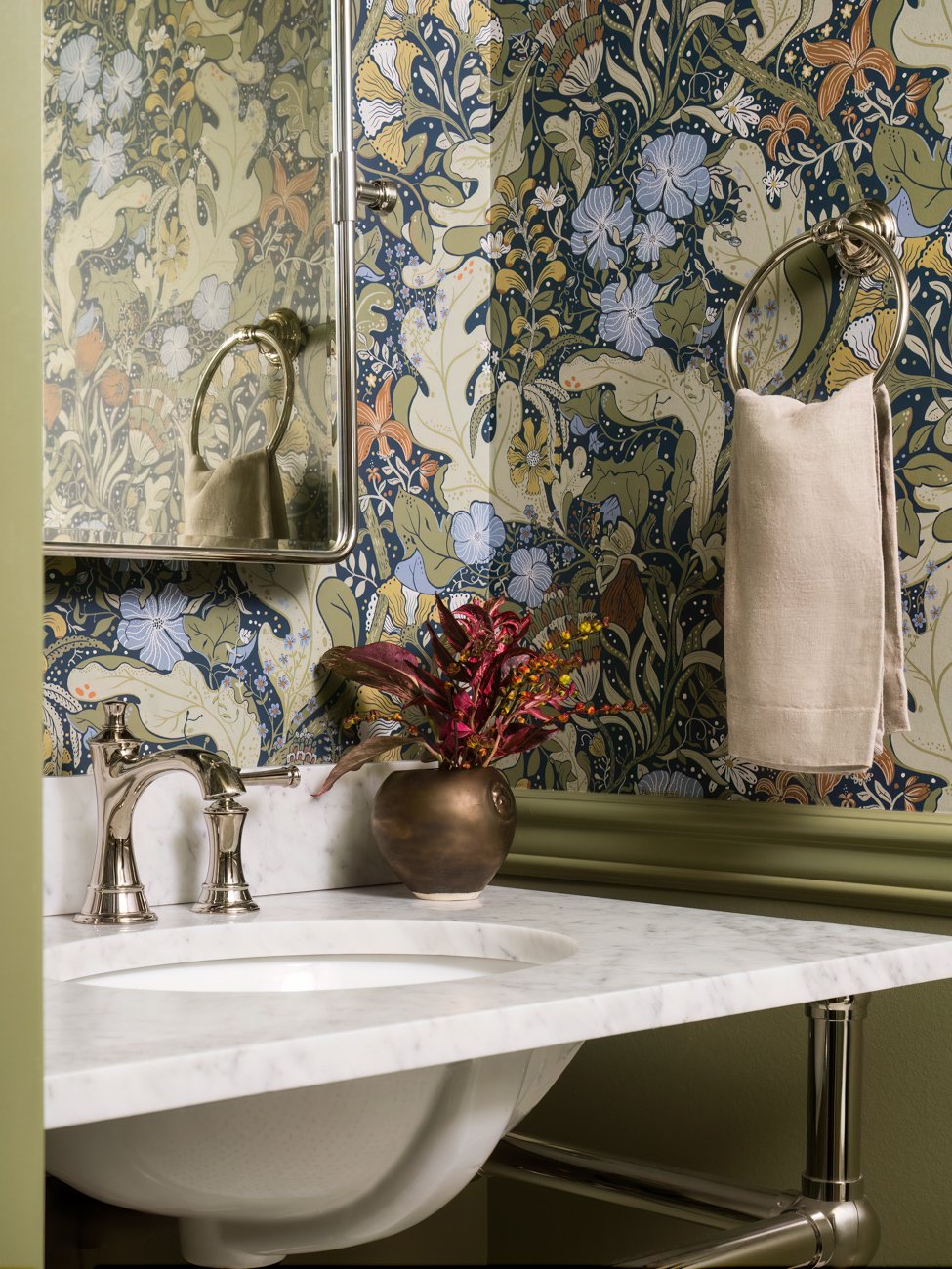 A bathroom with a distinctive floral wallpaper, a marble countertop with a white sink, a bronze vase with pink and green foliage, a towel on a ring, and a mirrored cabinet.
