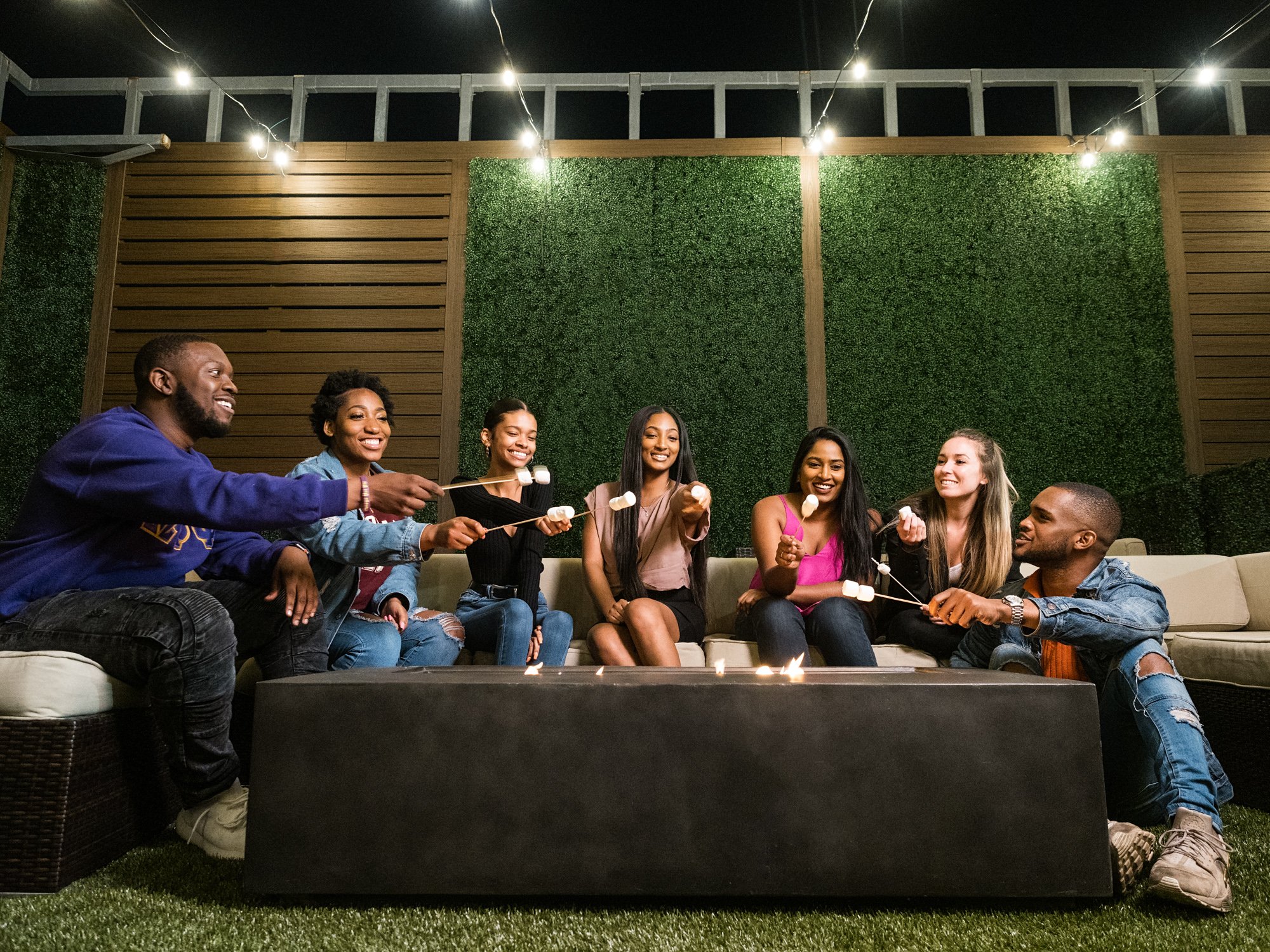A group of seven young adults happily roasting marshmallows on a bonfire in a backyard at night, with string lights hanging overhead and a wall of green foliage in the background.