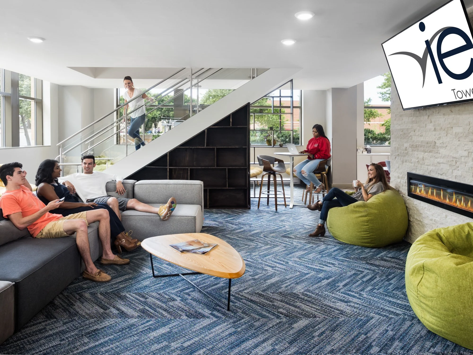 A modern lobby with multiple people relaxing and socializing, including young adults sitting on sofas and bean bags, a woman working on a laptop at a counter, and a woman climbing stairs, with a large window and a wall-mounted TV displaying a logo.