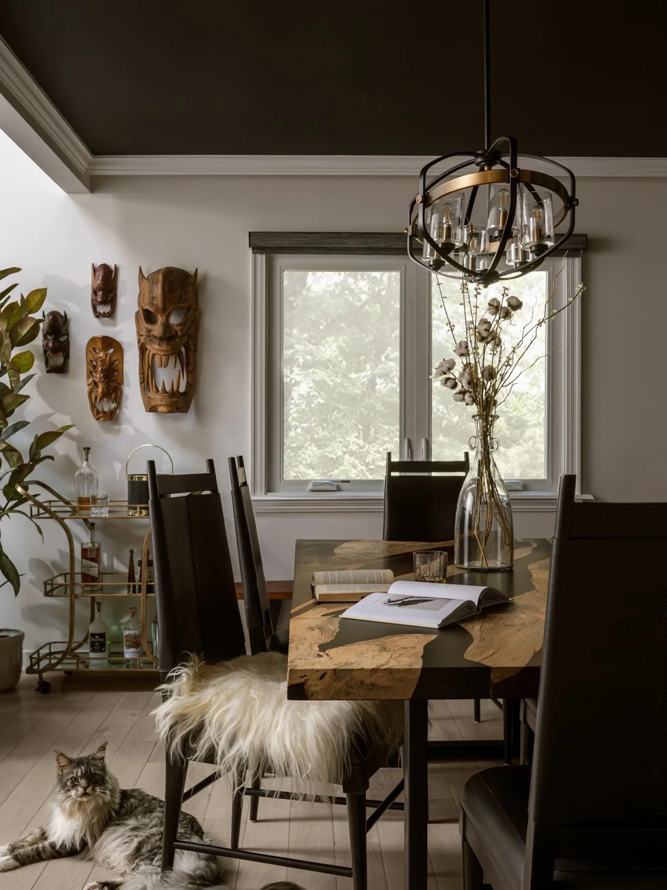 A dining room featuring a marble-top dining table with black chairs, a large window, a chandelier, wall masks, a glass vase with dried flowers, a bar cart with bottles, a potted plant, and a cat lying on the floor.