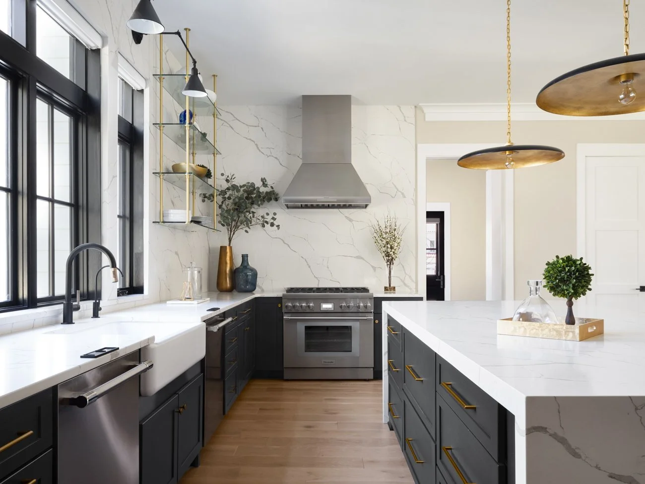 Modern kitchen with white marble countertops, black cabinets with gold handles, stainless steel oven, range hood, and black-framed windows. Decor includes potted plants, vases, and shelves with dishware. Wooden flooring and gold pendant lights.