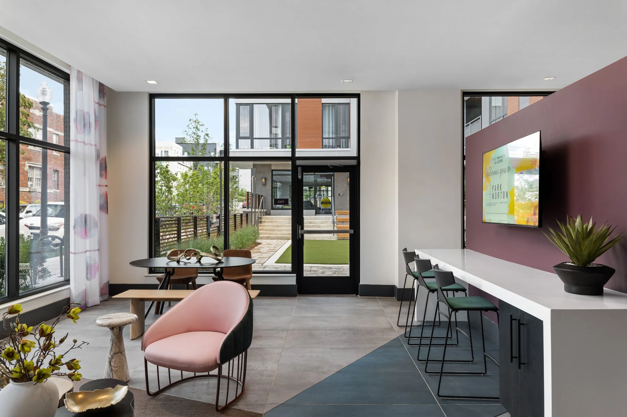 Modern lobby with large windows, sitting area with pink and black chairs, a white countertop with bar stools, and a purple accent wall with a mounted TV.
