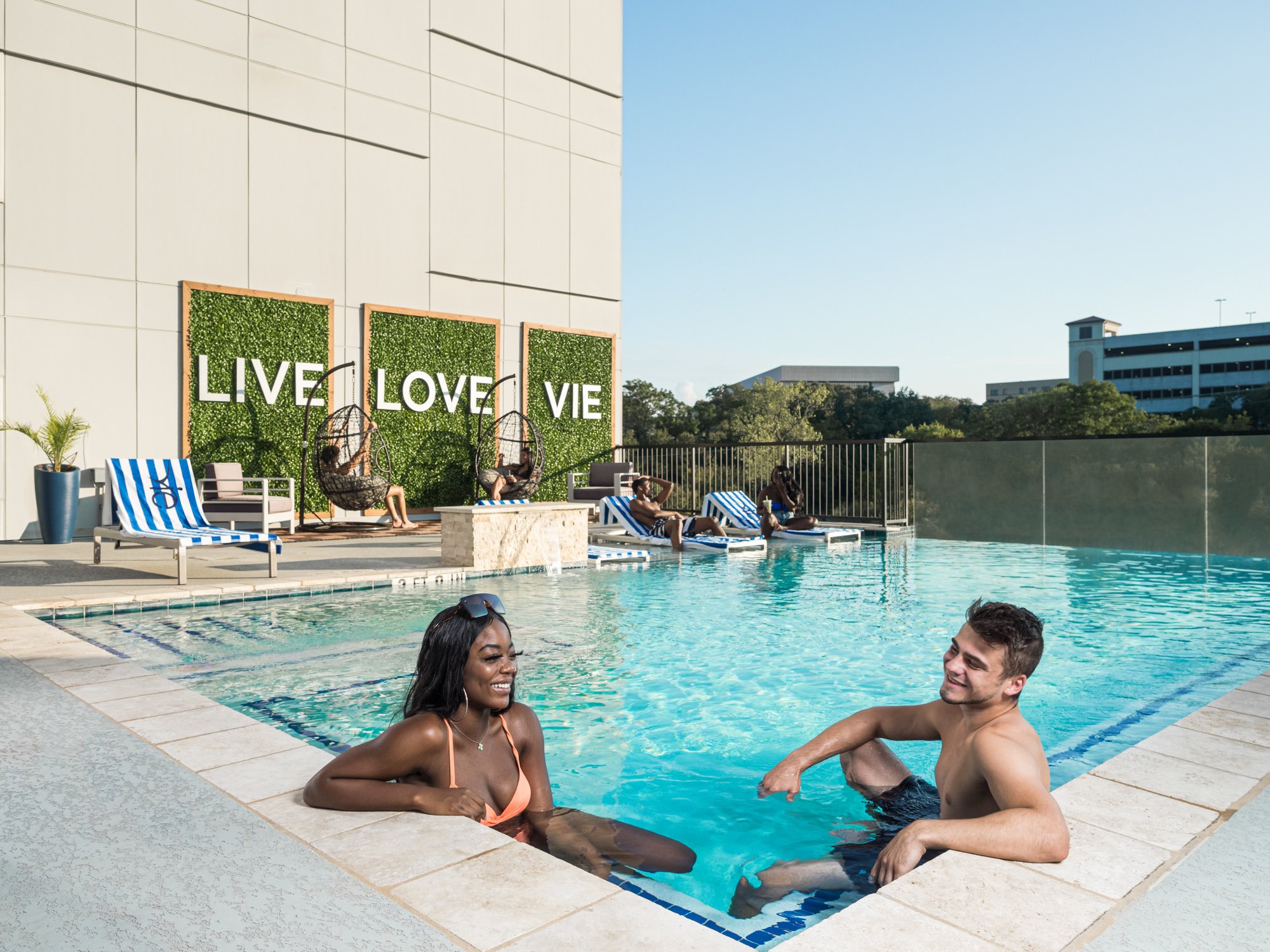 Two people relaxing in a rooftop swimming pool with a green wall that reads 'LIVE LOVE VIE' in the background, and other guests on lounge chairs and hanging chairs nearby.