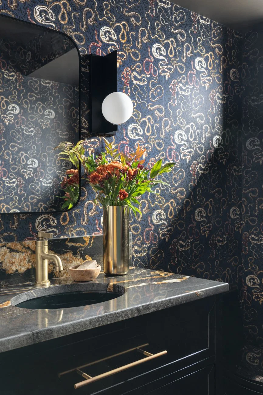 A bathroom vanity with a black cabinet, gold handle, and a dark gray marble countertop with gold veining. A gold vase with a colorful flower arrangement, a small stone bowl, and a gold and black wall sconce with a round white shade are on the counter