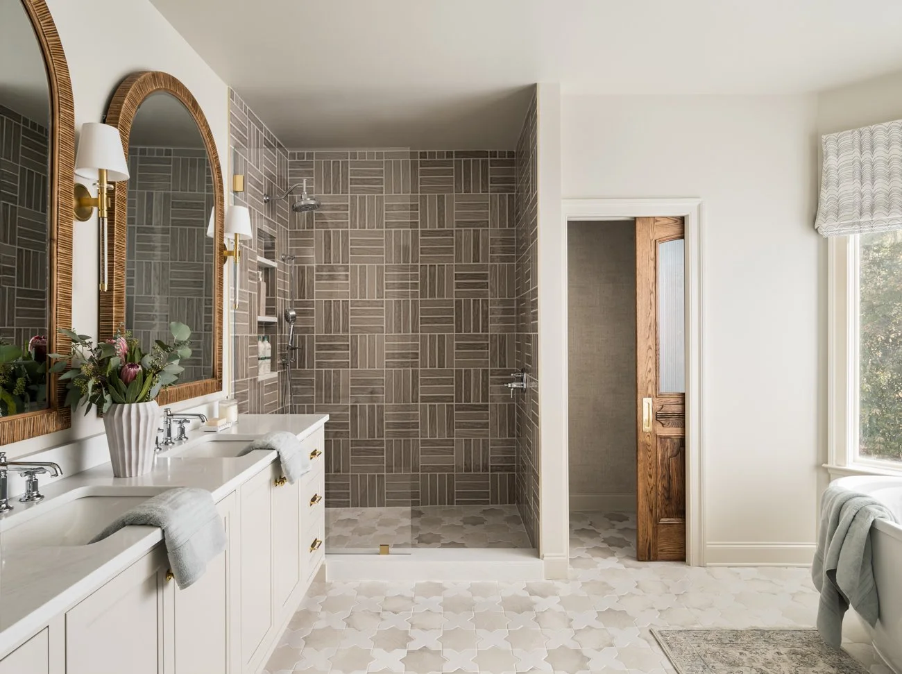 Bright bathroom with double vanity, large mirrors, floral arrangement, and walk-in shower with brown tiles and built-in shelves.