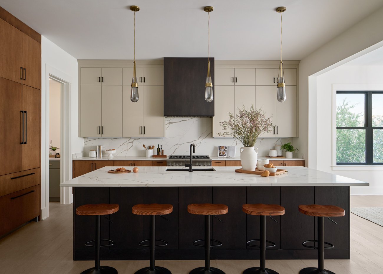 Modern kitchen with white marble island, black cabinets, beige and wood cabinetry, pendant lights, and large window showing greenery outside.