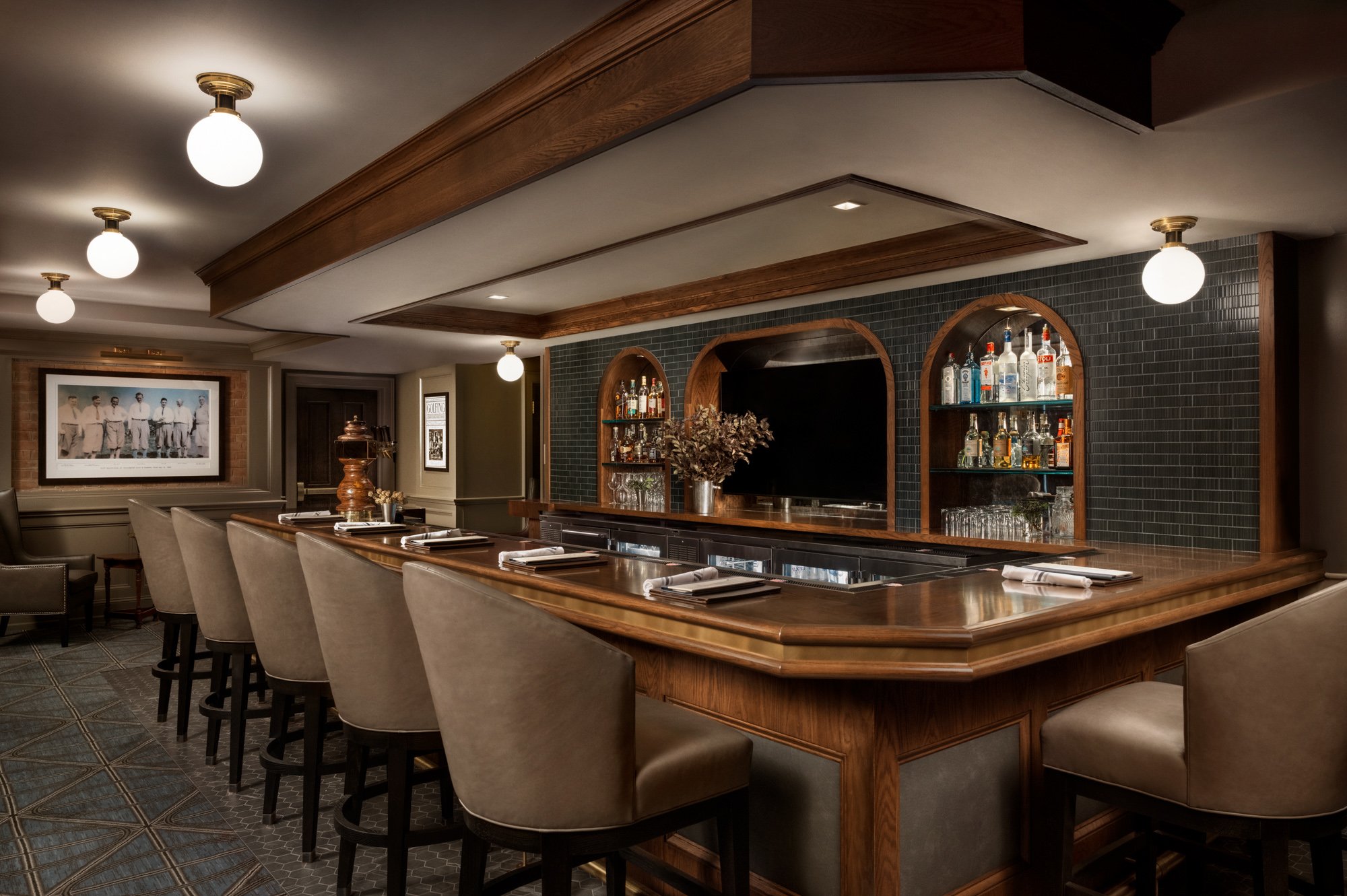 An interior shot of a bar with a wooden counter, bar stools, and a back wall with shelves of liquor bottles and glassware. There are ceiling lights and a framed photo on the wall.