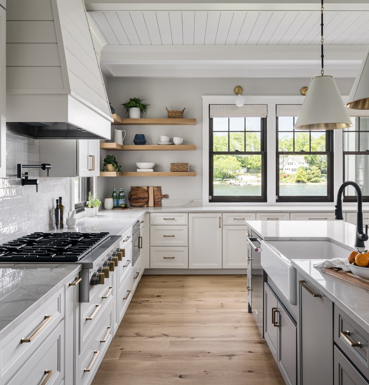 Modern kitchen with white cabinets, wooden open shelves, large windows overlooking a lake, and a kitchen island with a farmhouse sink.