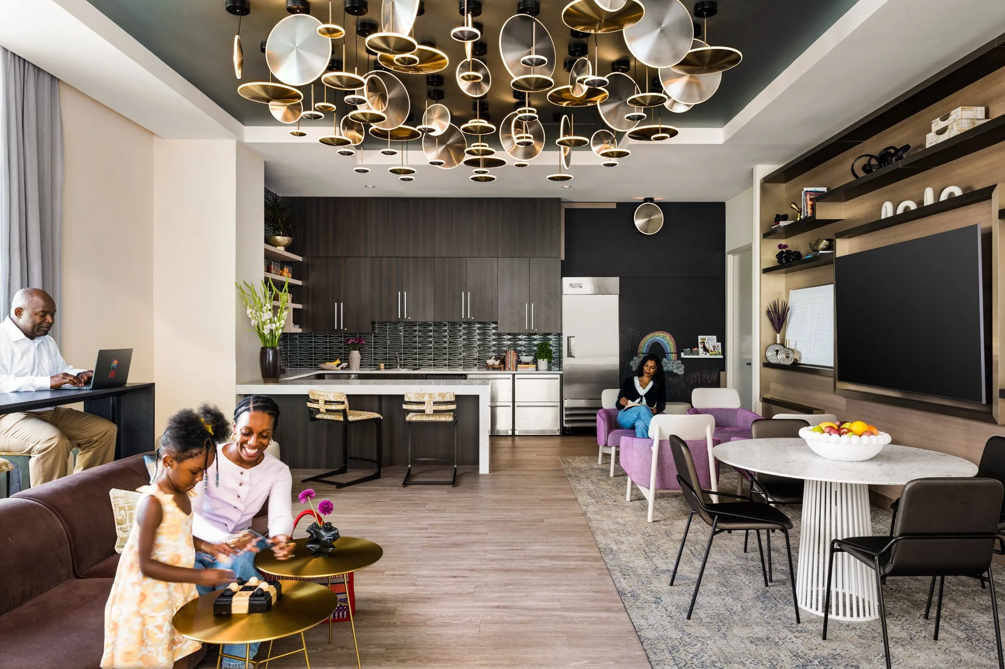 Modern living room with a kitchen in the background, featuring a large, artistic ceiling light fixture, a flat-screen TV, and several people, including a woman and girl playing a game on a small table, and a woman sitting on a purple armchair.