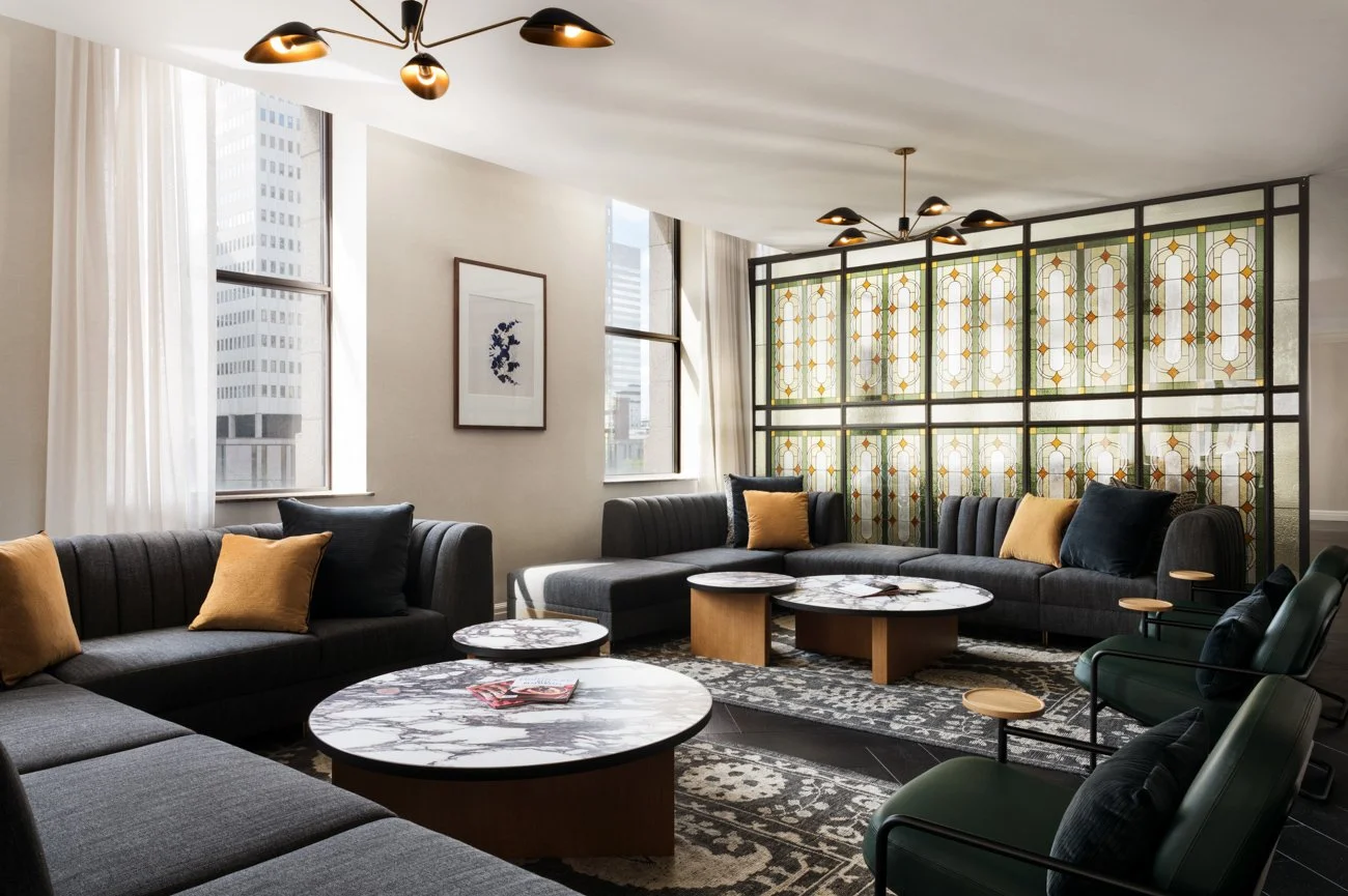 Modern living room with large windows, patterned stained-glass room divider, dark gray sofas with yellow and black pillows, marble-topped coffee tables, and green armchairs.