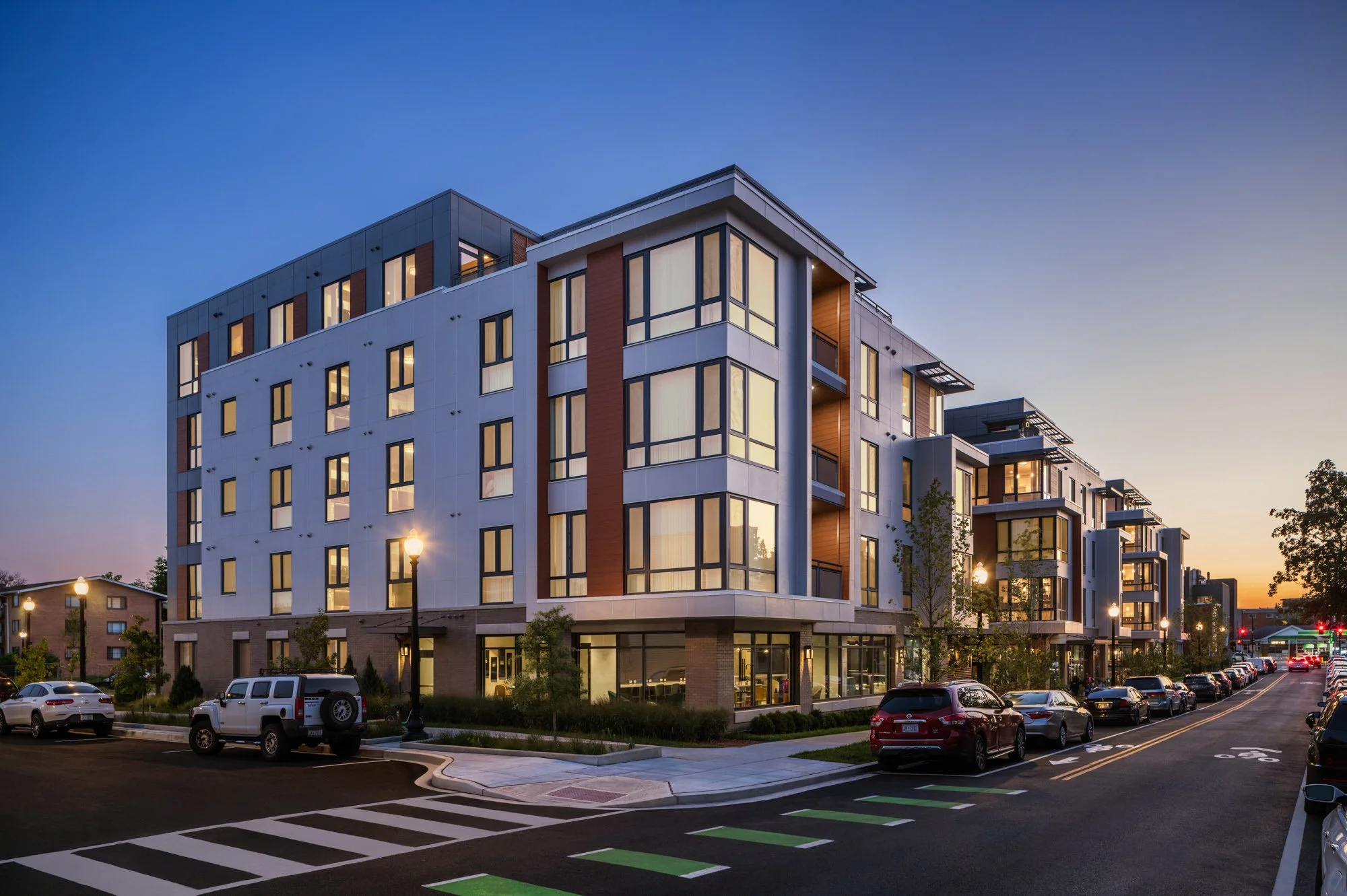 Modern multi-story apartment building with large windows, lit from within, at dusk. The street in front has parked cars and streetlights.