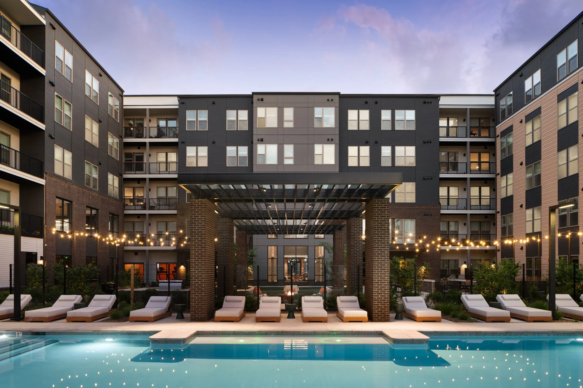 A modern apartment complex with a swimming pool, lounge chairs, string lights, and a brick and dark-colored building facade during dusk.
