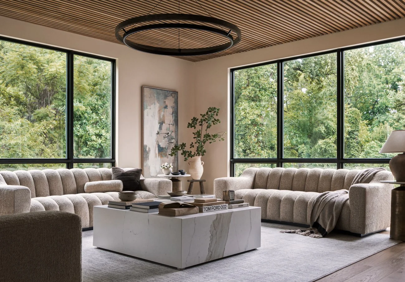 Modern living room with large windows, beige sofas, a white marble coffee table, and decorative items including a large vase with greenery, books, and artwork on the wall.