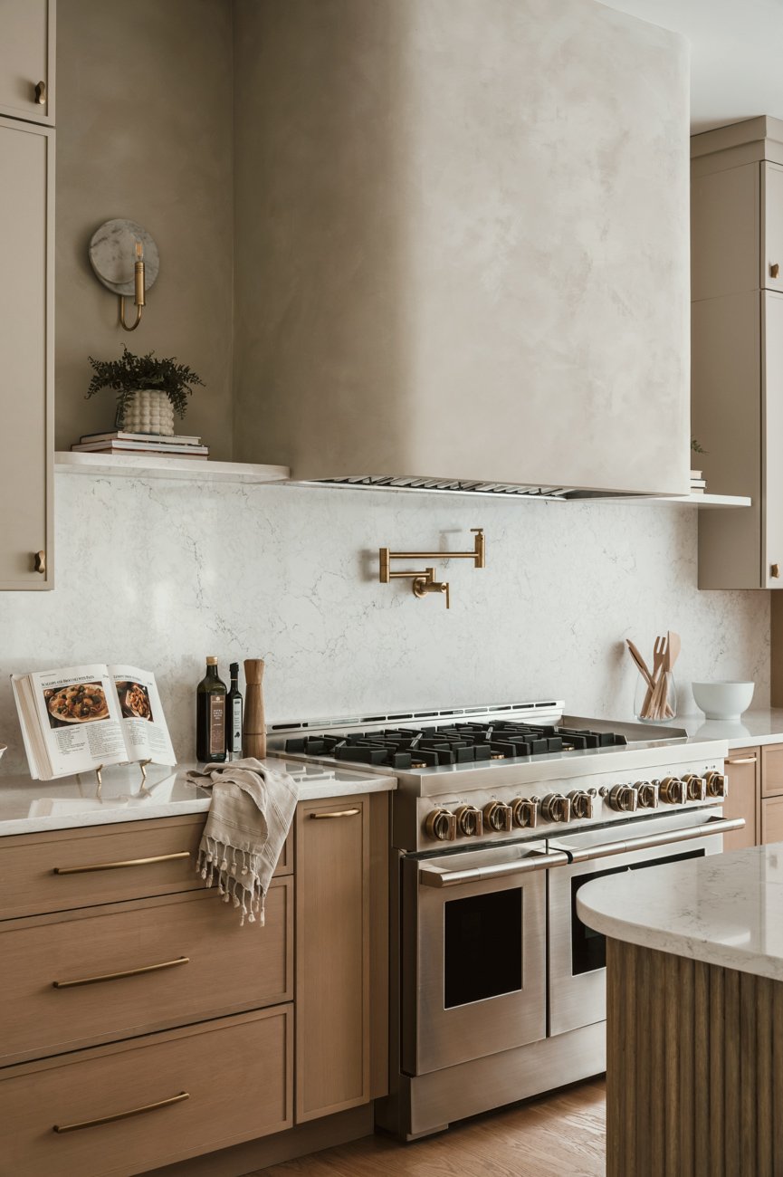 Modern kitchen with beige cabinetry, a marble backsplash, and a stainless steel oven and stovetop. Decor includes a cookbook on a stand, bottles, a bowl, and utensils.