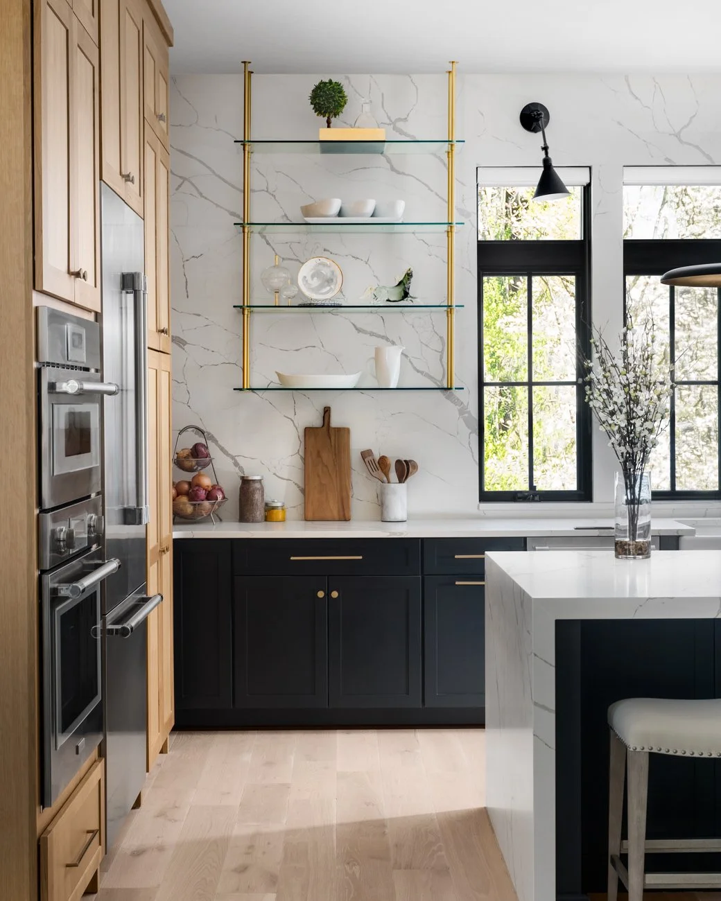 Modern kitchen with wooden cabinets, black lower cabinets, marble countertops, open shelving with decor, large windows, and a vase with white flowers.