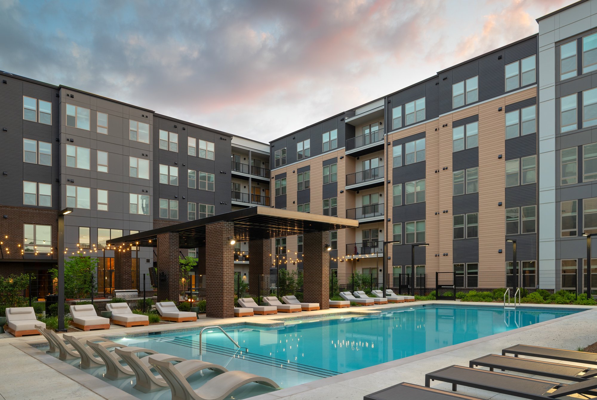 Outdoor swimming pool area in an apartment complex during evening, with lounge chairs, string lights, and modern apartment buildings in the background.