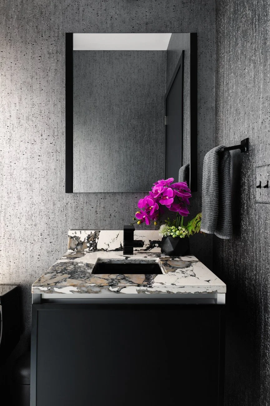 Modern bathroom with gray textured walls, a large mirror, a black sink faucet, a marble countertop with black and white patterns, a black vase with purple flowers and greenery, and gray towels on a black towel rack.
