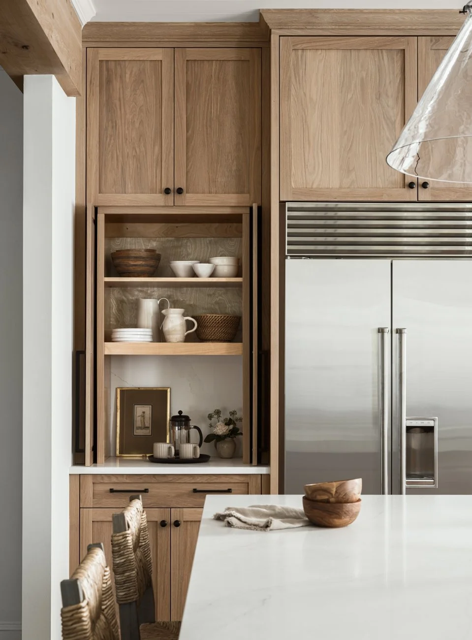 A kitchen with wooden cabinets, open shelves displaying dishes and bowls, a stainless steel refrigerator, a white countertop with bowls and a napkin, and woven barstools.