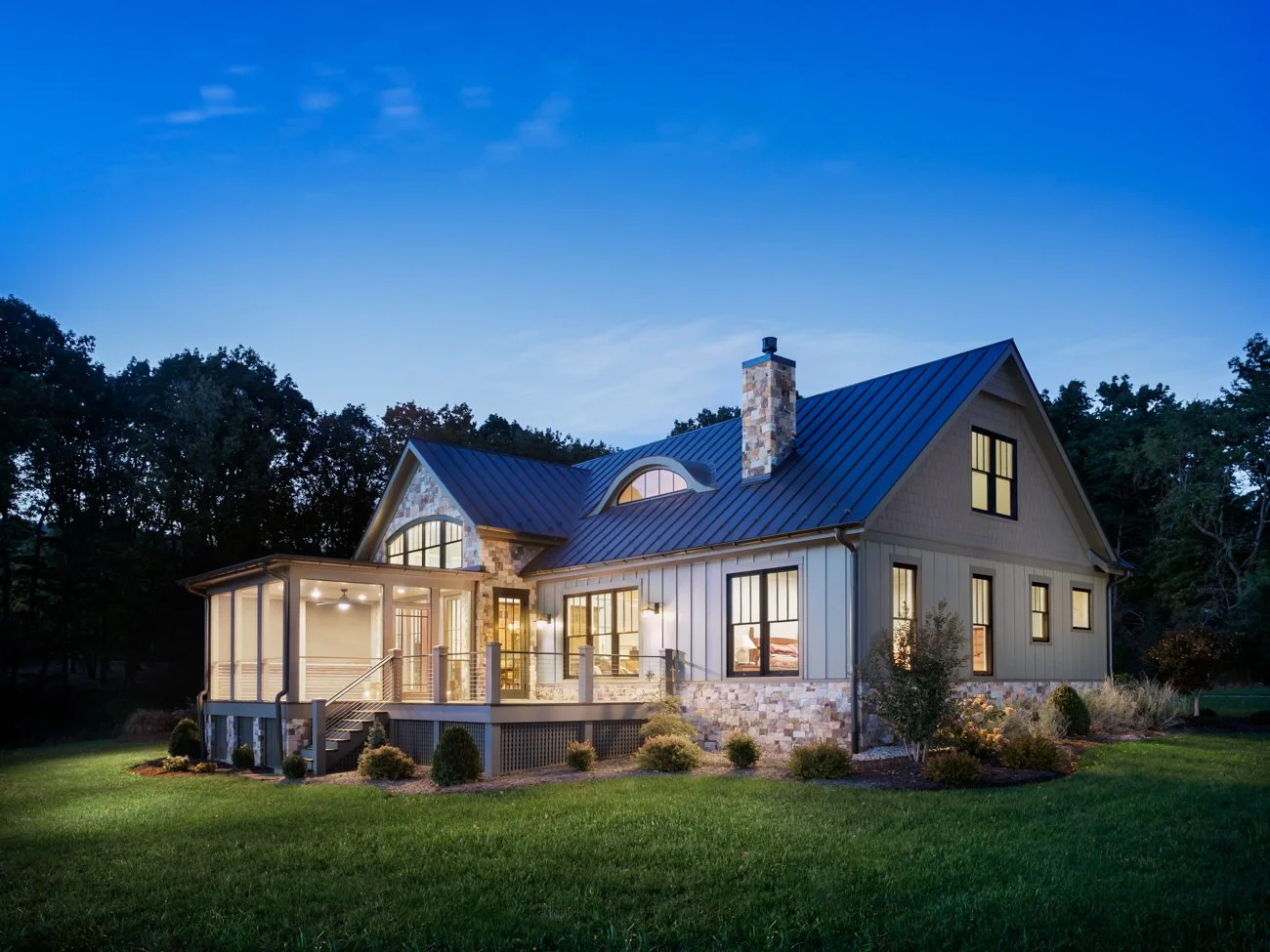 A modern house illuminated at dusk with large windows, a stone chimney, and a spacious porch, surrounded by a well-maintained lawn and trees.