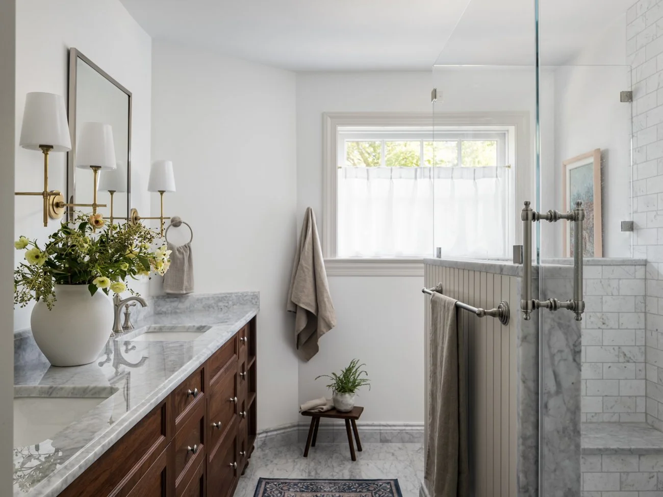 Bright bathroom with a marble countertop vanity, wooden cabinets, a large mirror, a floral arrangement, and a walk-in shower with white tiles and a glass door. Natural light from a window.