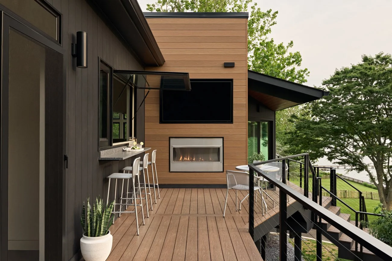 View of an outdoor balcony of a modern house with wood and dark-colored siding, outdoor fireplace, TV, and seating area with tables and chairs, surrounded by trees and greenery.