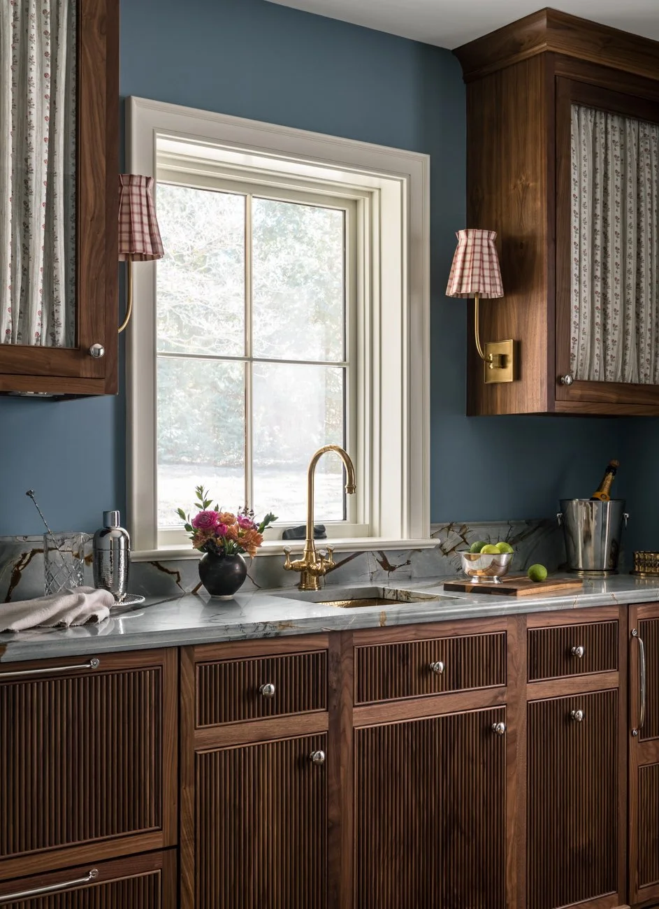 A kitchen with a window above a marble countertop, wooden cabinets with fluted panels, a brass faucet, and wall-mounted lamps with plaid shades. Decorations include a vase of flowers, a bowl of green apples, and a champagne bucket.