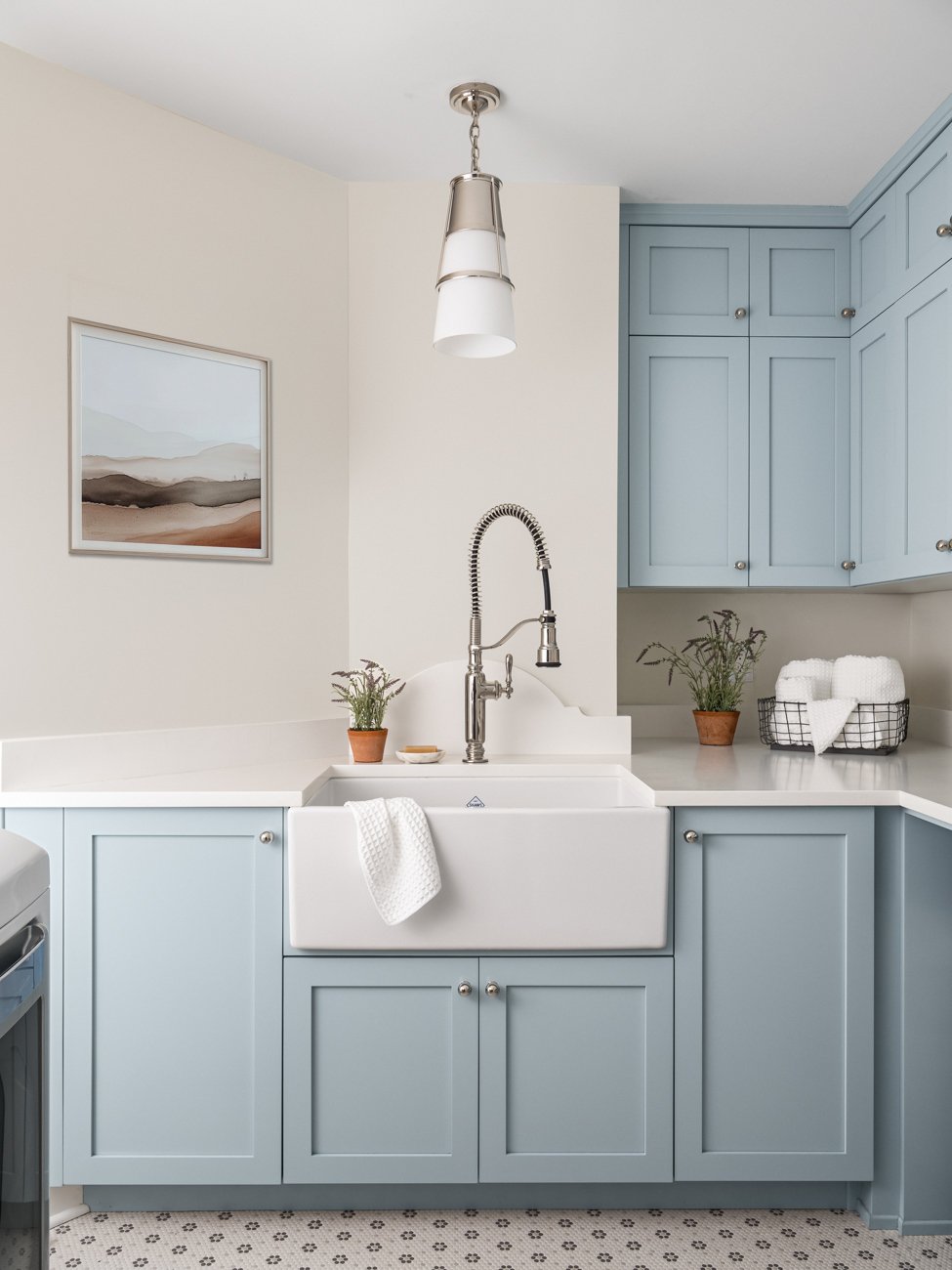 Kitchen with light blue cabinets, a white farmhouse sink, and a modern pendant light. Small potted plants and towels are on the counter.