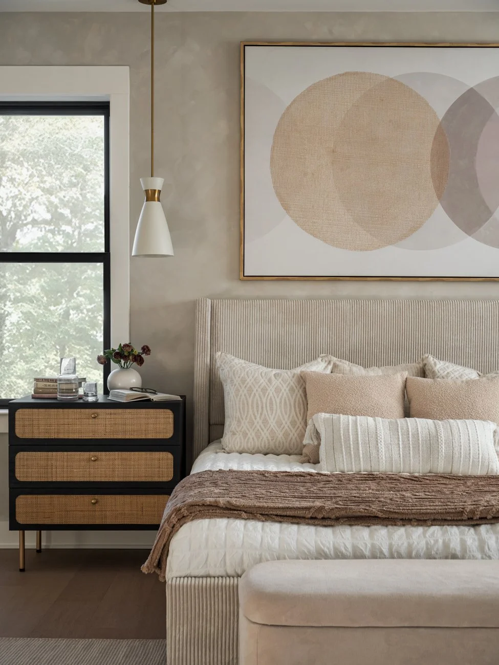 A neatly made bed in a bedroom with a plush headboard, decorative pillows, and a brown throw blanket. Next to the bed is a black dresser with rattan drawer fronts, a white vase with flowers, books, and glasses. A large abstract art piece with circles
