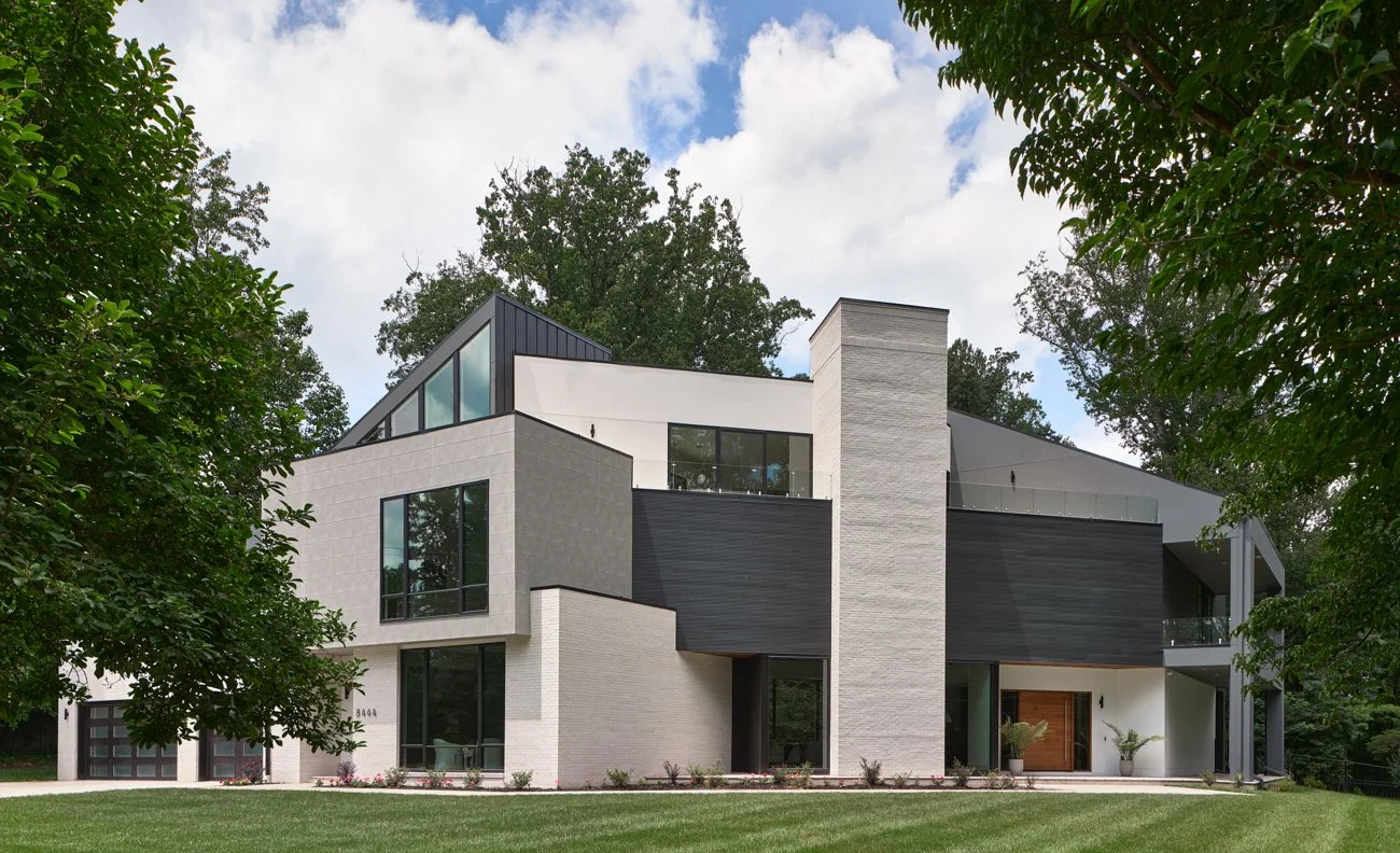 Modern multi-story house with large windows, white and black exterior, surrounded by greenery, under a partly cloudy sky.