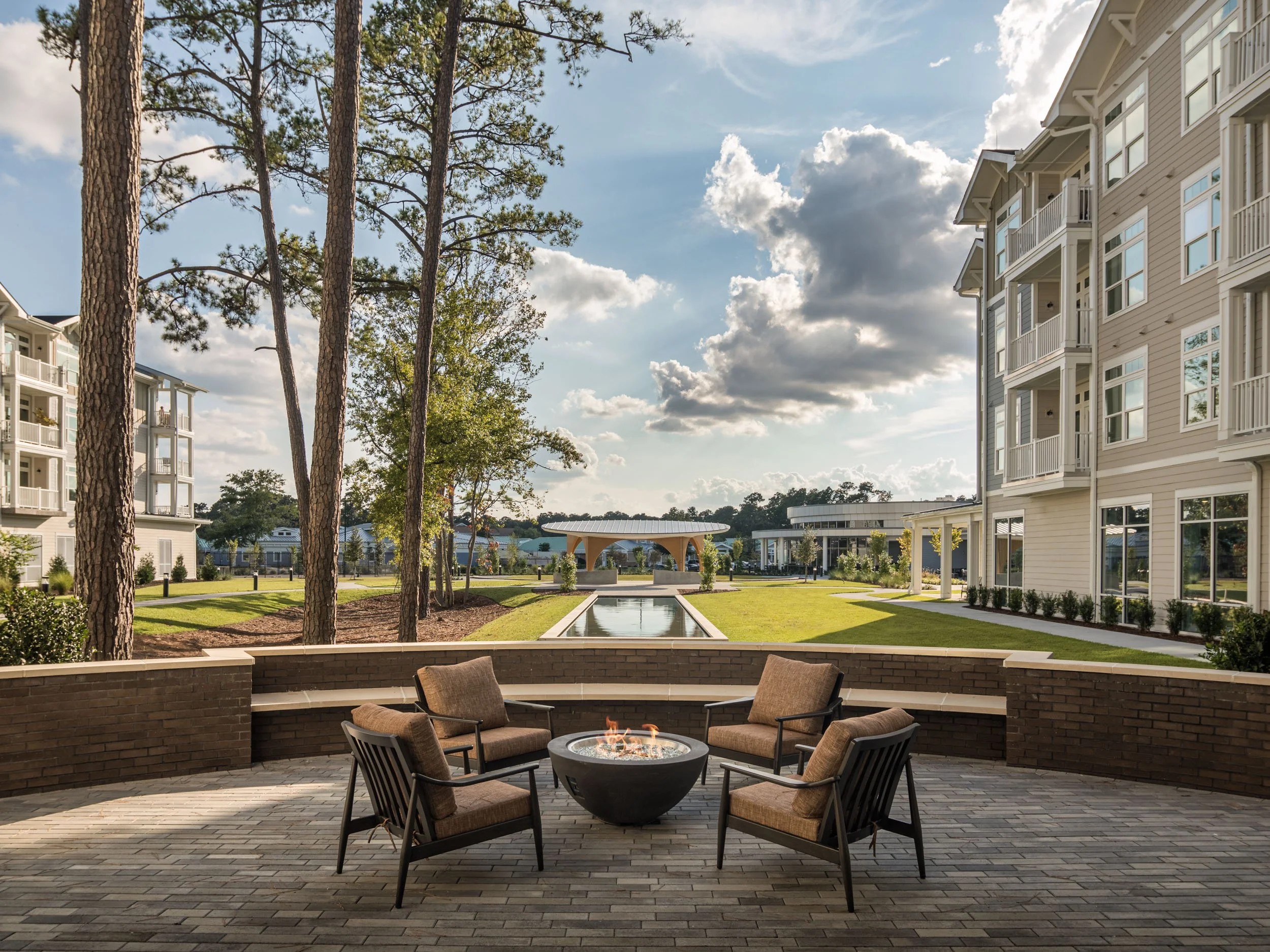 Outdoor patio with four chairs around a fire pit, overlooking a landscaped courtyard with a fountain, tall trees, modern residential buildings, and a pavilion in the background under a partly cloudy sky.